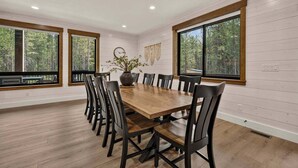 A dining room with a wooden table and chairs, large windows with a view of trees, and a vase with flowers on the table. - Pine and Sky Lodge+WiFi+AC+Hotub+30Min2Yellowstone (Island Park)