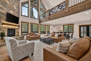 A spacious living area with a stone fireplace, multiple seating arrangements, and large windows offering views of the outdoors. - Pine and Sky Lodge+WiFi+AC+Hotub+30Min2Yellowstone (Island Park)
