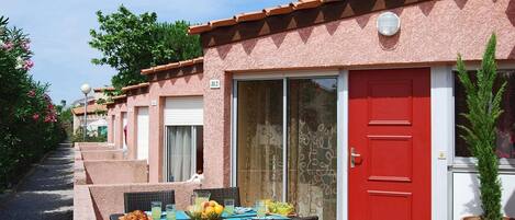 A row of rooms with a red door, a glass door, and a balcony with wicker furniture and a fruit bowl.
