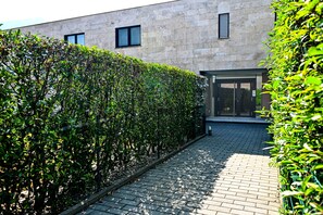 Ein gepflasterter Weg, gesäumt von ordentlich geschnittenen Hecken, führt zu einem Gebäude mit moderner Architektur und einem Glaszugang.