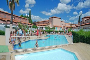 A swimming pool area with a fenced pool, palm trees, and buildings with red-tiled roofs.