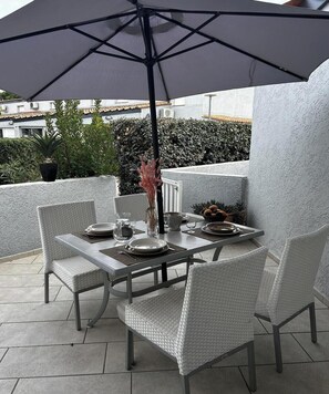 A table set outdoors with a large umbrella, surrounded by wicker chairs, on a tiled patio with a view of a building and greenery.