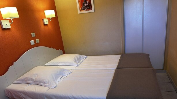 Uma cama de casal com roupa de cama branca e marrom, duas luminárias fixadas na parede, uma pequena estatueta e uma porta fechada.