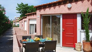 Uma fileira de quartos, com porta vermelha, porta de vidro e varanda com móveis de vime e uma fruteira.