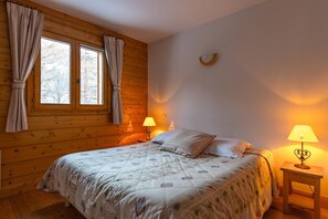 A bedroom with a wooden ceiling, a bed with a floral bedspread, two bedside lamps, and a window with curtains.