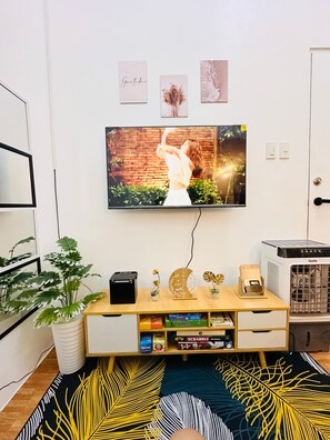 A living room with a flat-screen TV mounted on the wall, a wooden entertainment unit with a few decorative items, a potted plant, and a patterned rug.