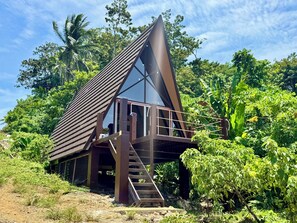 A modern, triangular-roofed structure with a staircase leading to a balcony, surrounded by lush greenery.