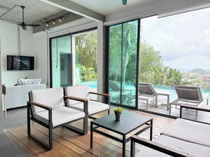 Ein modernes Wohnzimmer mit einer Couch, Sesseln und einem Couchtisch, einem an der Wand montierten Fernseher sowie einer großen Schiebeglasdoor, die zu einem Außenbereich mit Pool und Liegestühlen führt.
