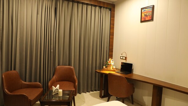 Habitación de hotel con un ventanal grande, una cama, un escritorio con una computadora portátil, dos sillas y una pequeña mesa con una vela y un objeto decorativo.