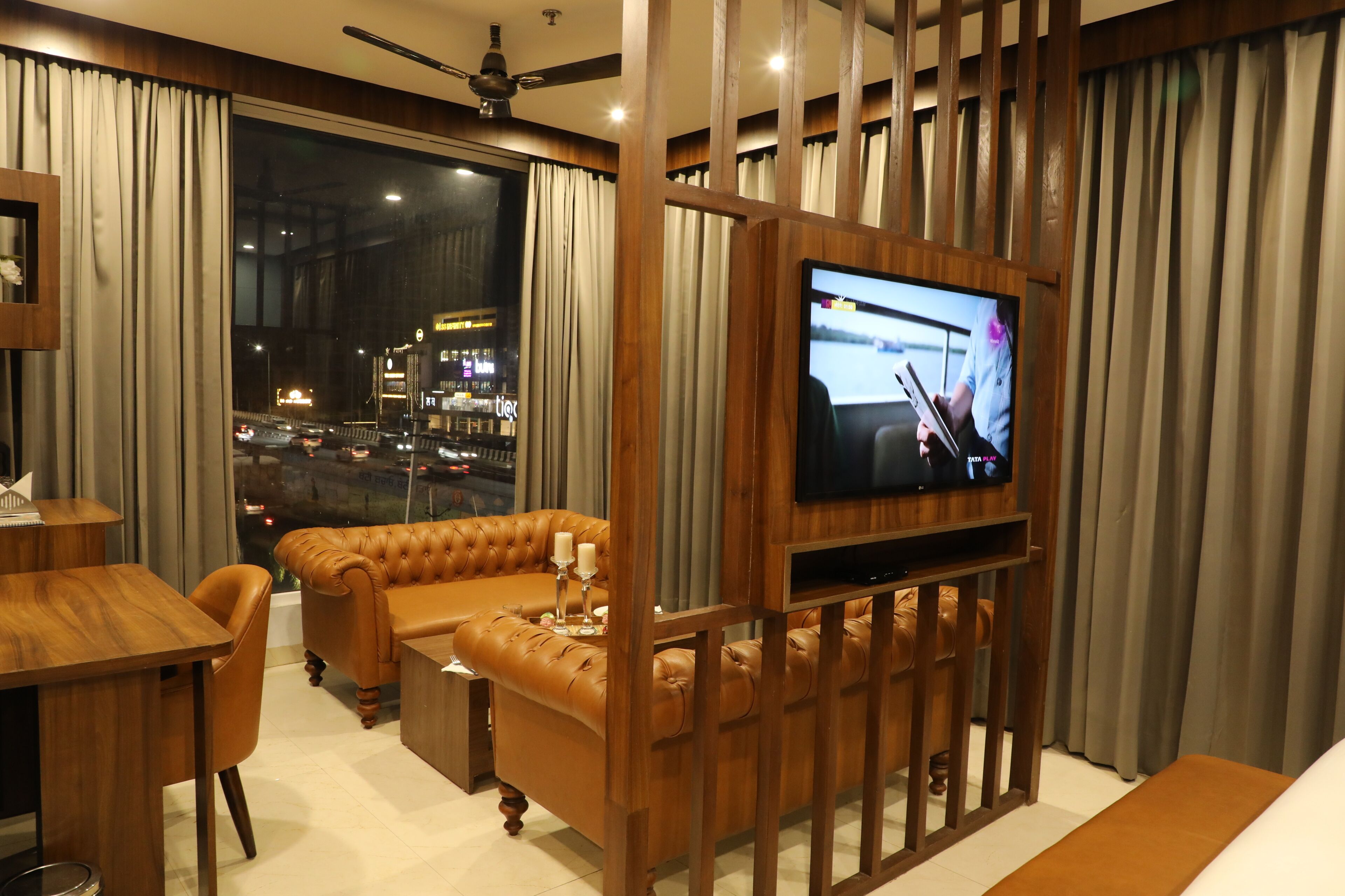 A modern living room with a large flat-screen TV, leather sofas, a wooden coffee table, and a view of a cityscape through large windows.