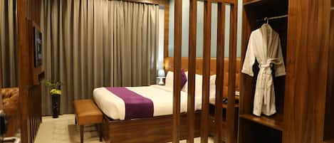 A hotel room with a bed, a robe, a wooden wardrobe, a bench, and a small table with a lamp.