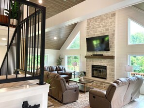 A modern living room with a stone fireplace, leather sofas, a flat-screen TV, and a wooden staircase. - Wooded Family Escape | Grill | DiscGolf | Big Yard (Plymouth)
