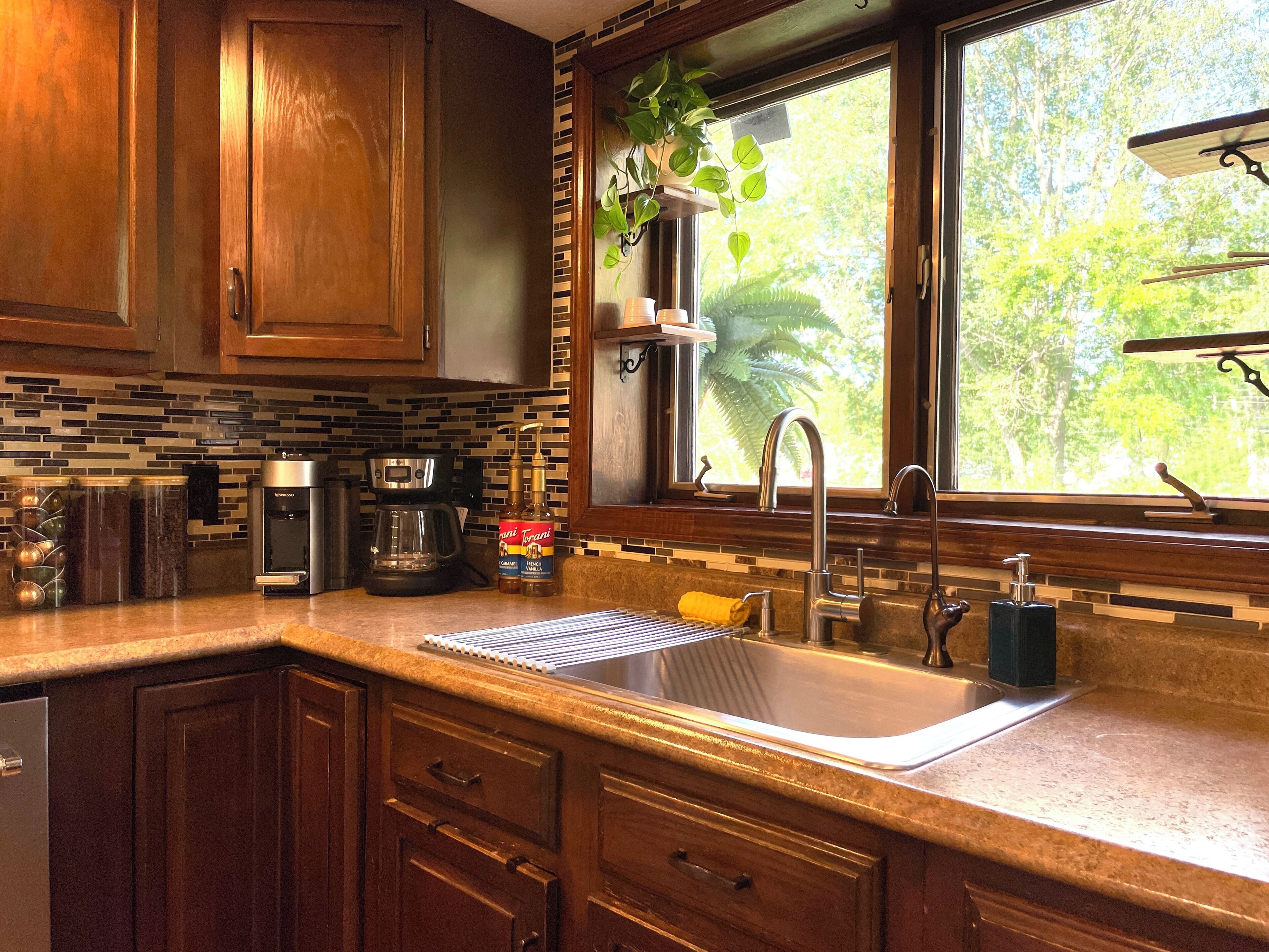 Una cocina con armarios de madera, un lavamanos de acero inoxidable, una cafetera y una ventana con vistas a árboles.