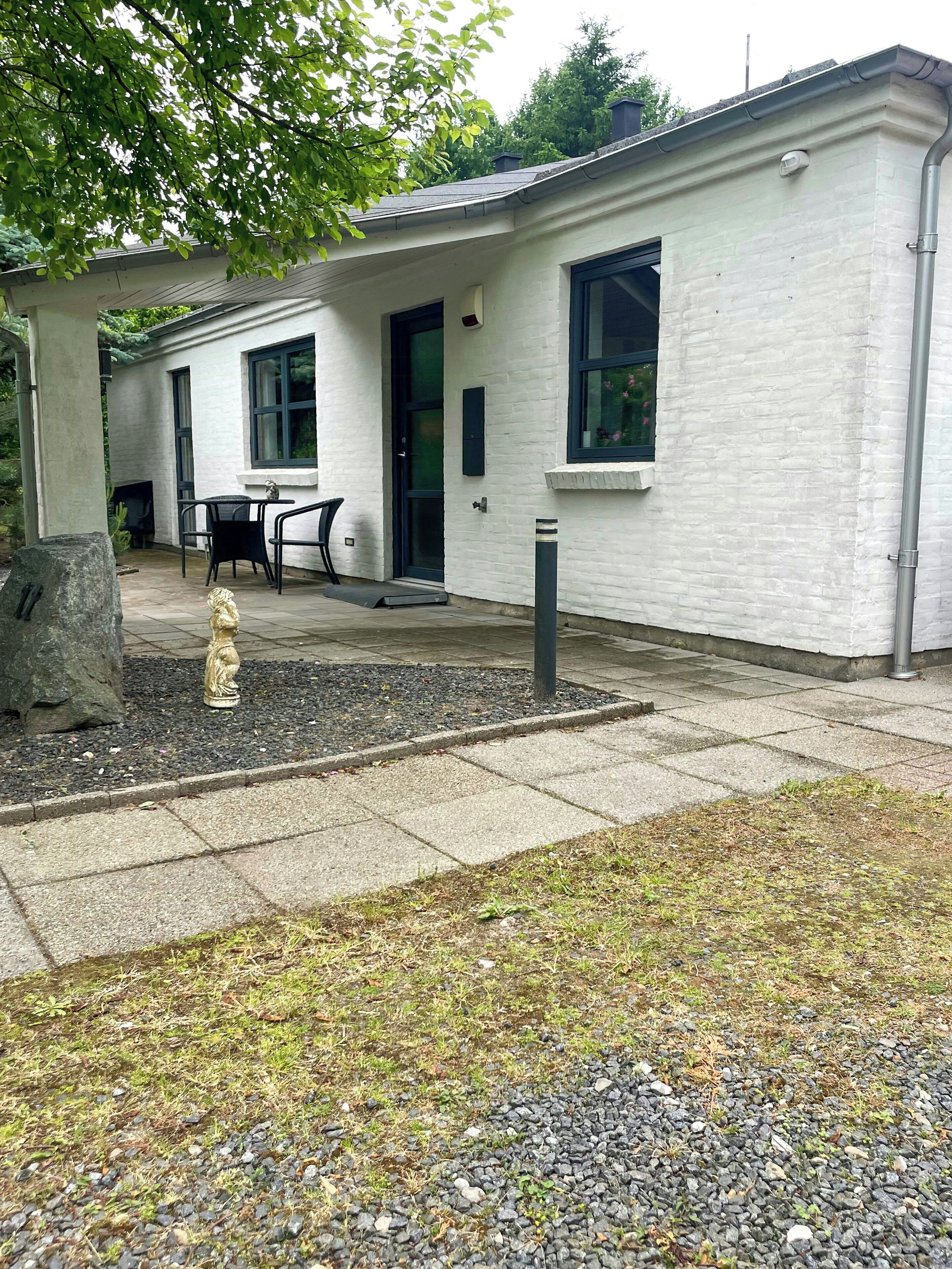 A building with a white exterior, a black door, and a window. There is a patio area with a table and chairs, and a statue in the foreground.