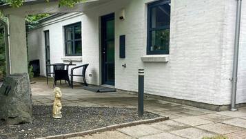 A building with a white exterior, a black door, and a window. There is a patio area with a table and chairs, and a statue in the foreground.