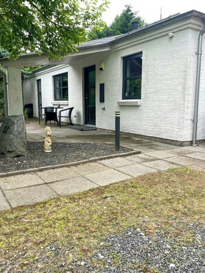 Edificio con fachada blanca, puerta negra y una ventana. Se observa un patio con una mesa y sillas, además de una estatua al frente.