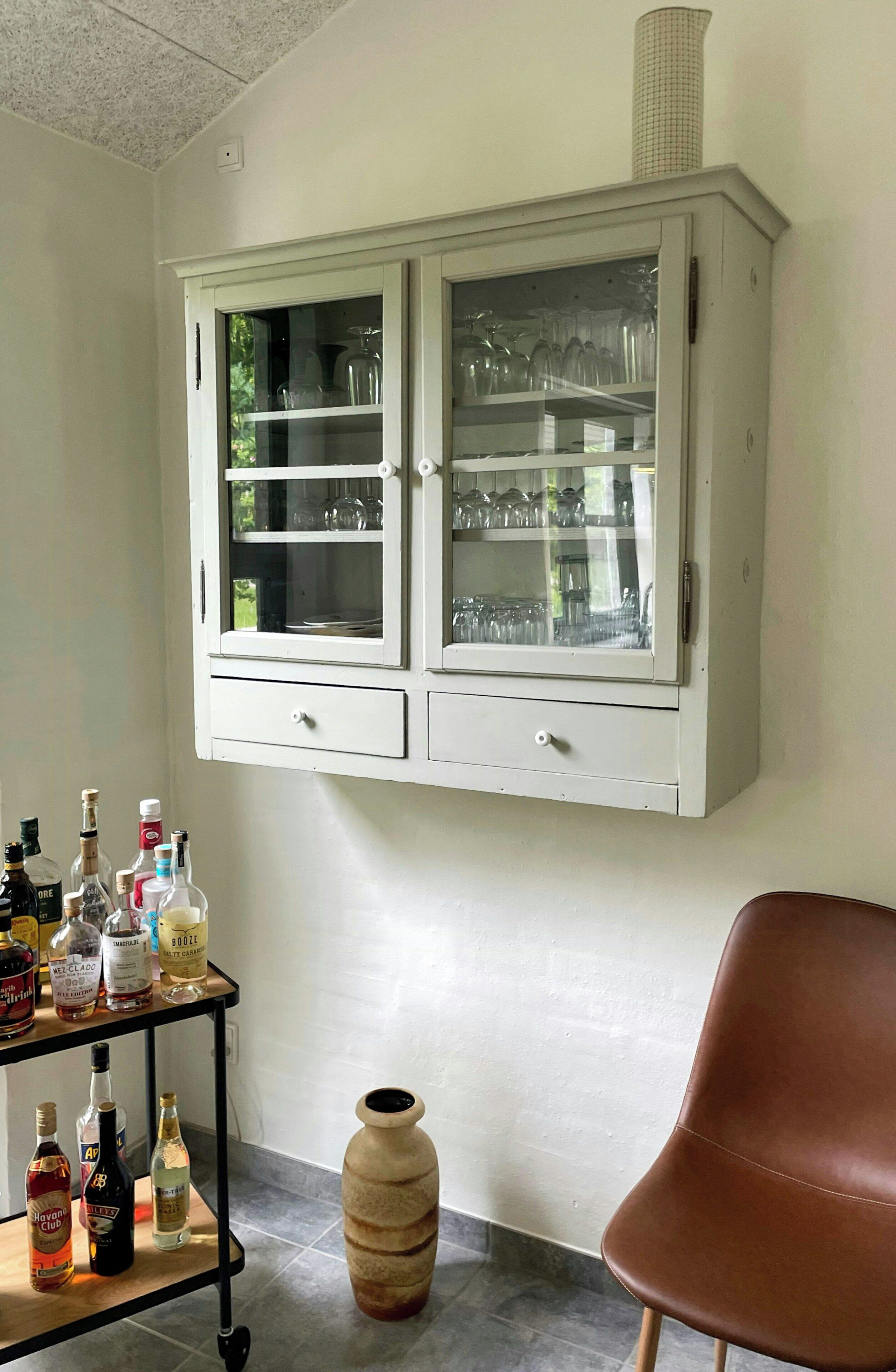 Ett rum med ett vitt skåp som innehåller glasvaror, ett litet bord med flaskor med sprit och en brun stol.