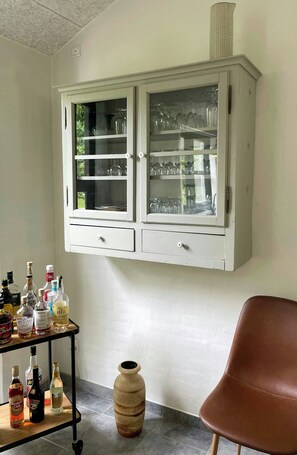 A room with a white cabinet containing glassware, a small table with liquor bottles, and a brown chair.