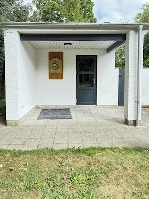 A small building with a dark door, a wooden sign above the door, and a black mat at the entrance.