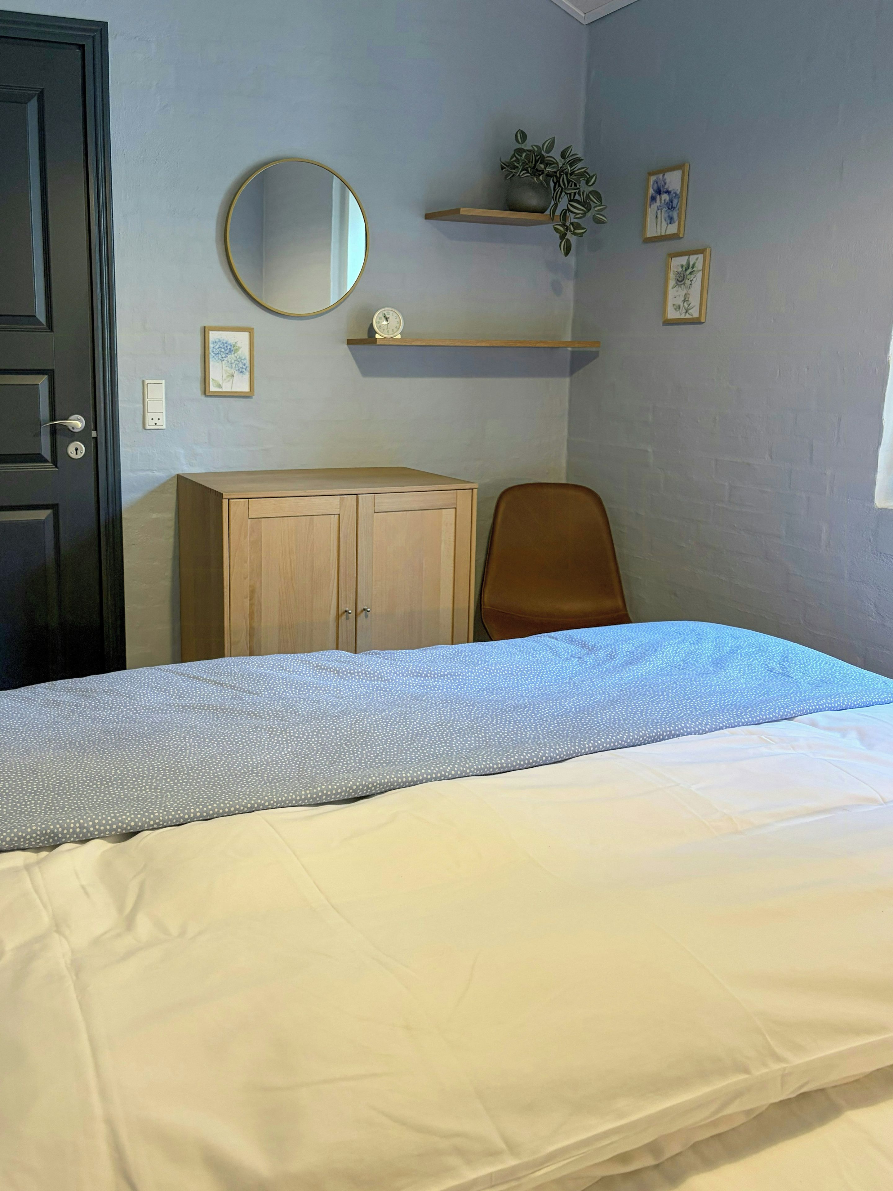 A bedroom with a bed, a wooden wardrobe, a chair, a mirror, and a shelf with a plant and pictures.