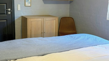 A bedroom with a bed, a wooden wardrobe, a chair, a mirror, and a shelf with a plant and pictures.