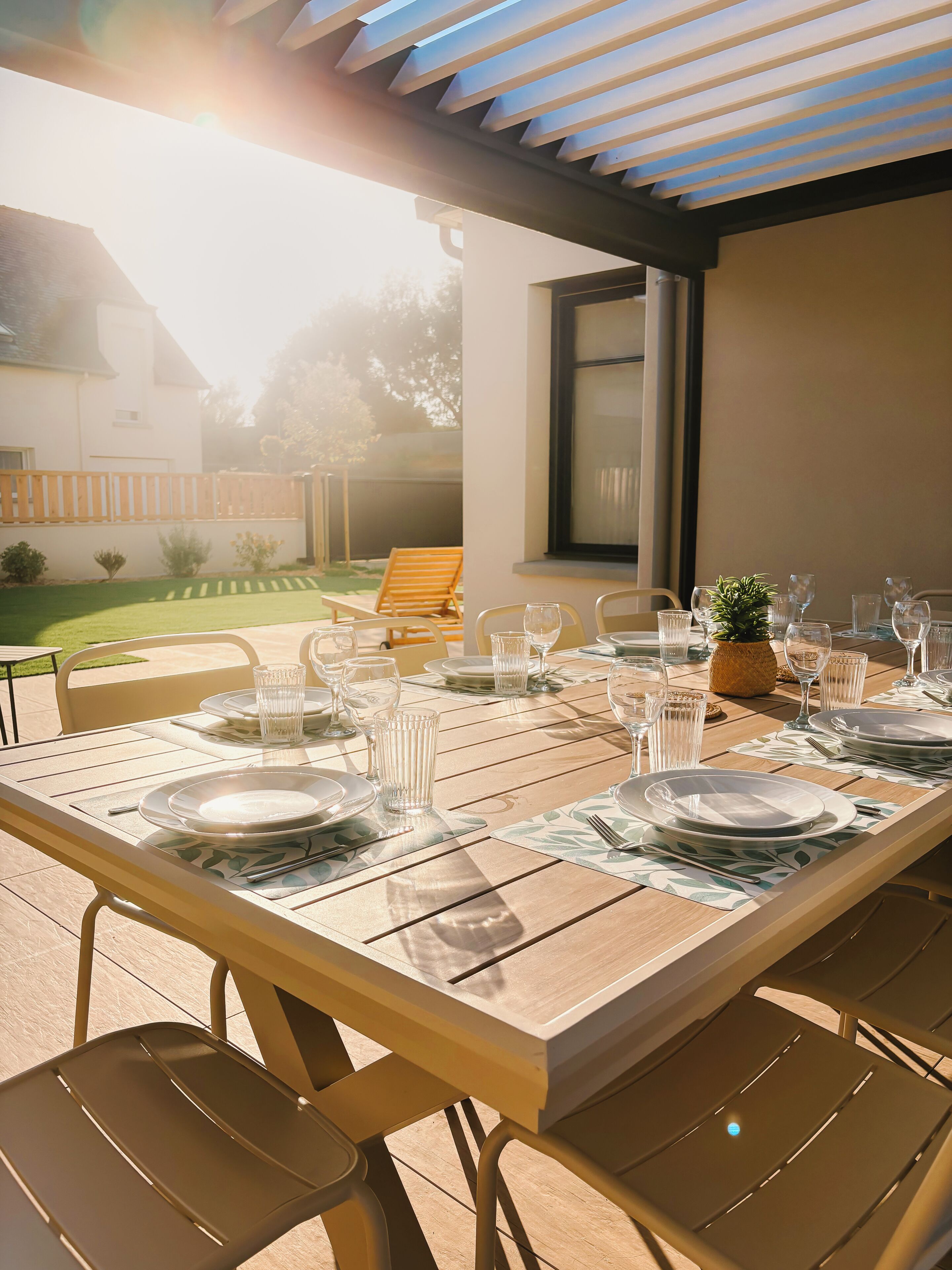 Un área de comedor al aire libre con una mesa lista para cuatro personas, rodeada de sillas, bajo una pérgola.
