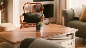 Ein Wohnzimmer mit Holztisch, Korbstuhl, Sofa, Stehlampe und einem Fenster mit Vorhängen.