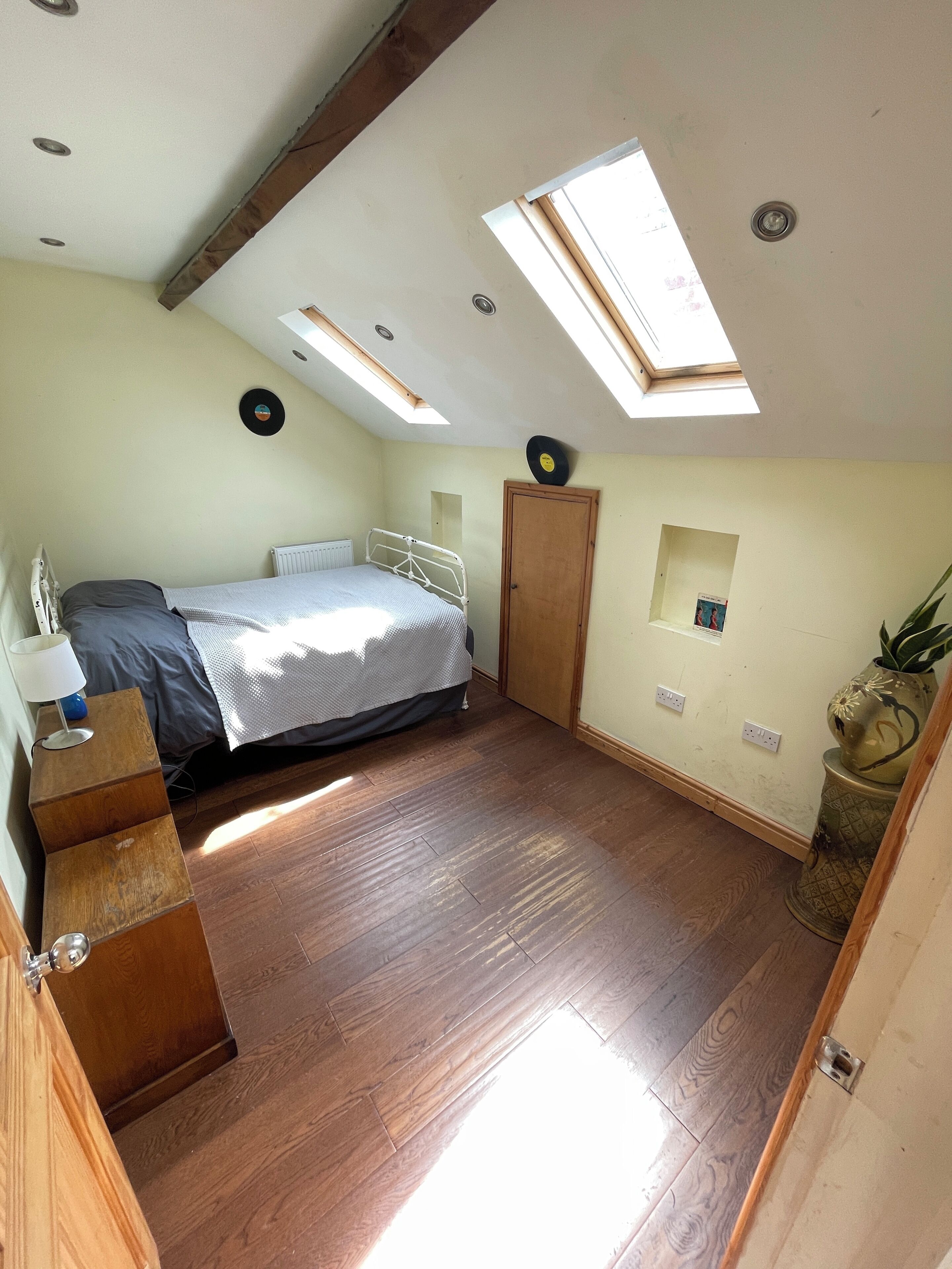 Een slaapkamer met een bed, dakramen, een houten dressoir en een klein plankje met een vaas.