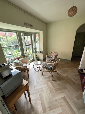 A room with a herringbone floor, wicker furniture, a table with a printer, and a view of a garden through large windows.
