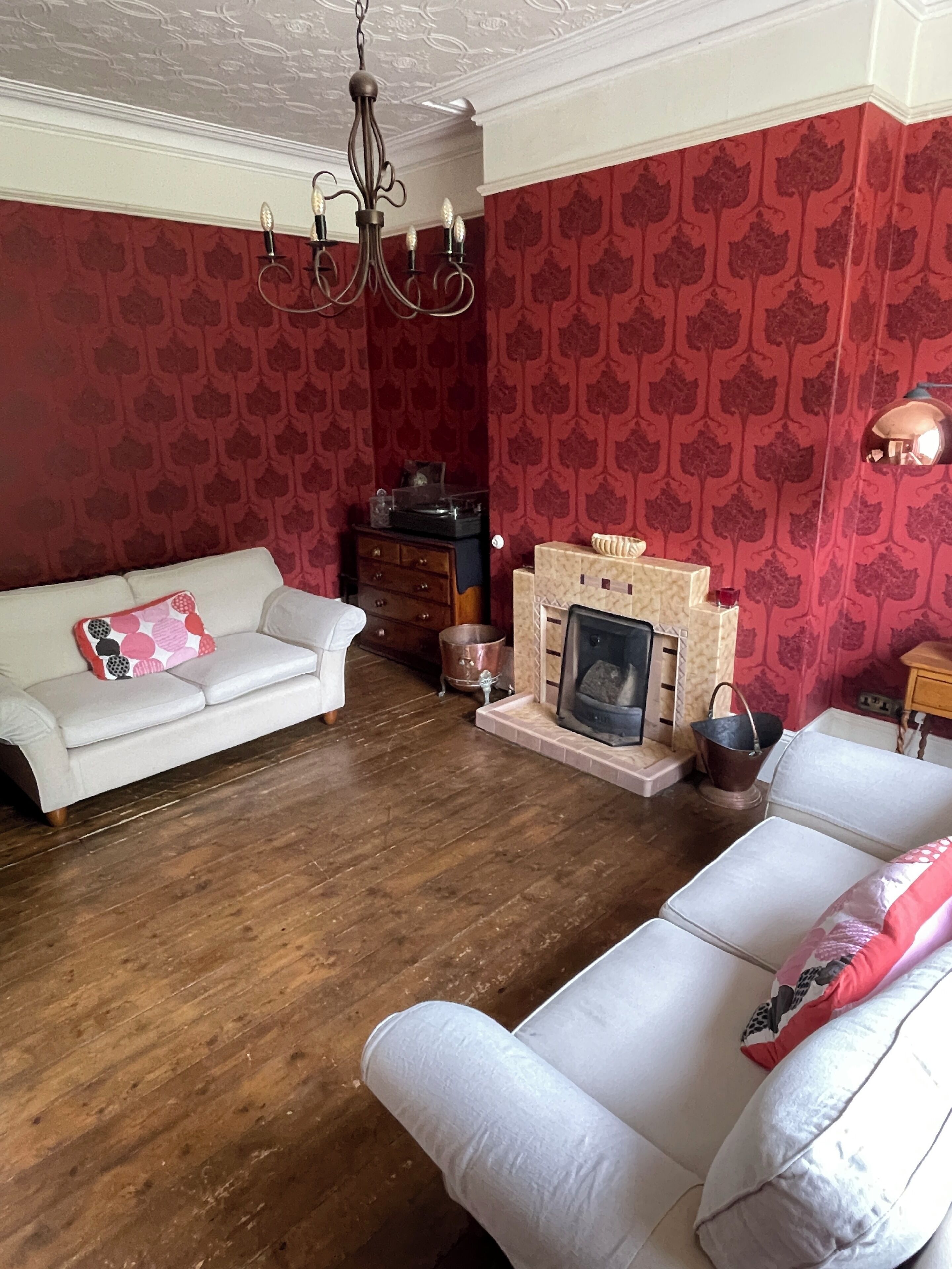 A living room with a fireplace, two white sofas, a wooden floor, and red patterned wallpaper.