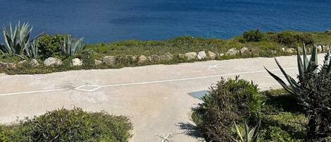 A paved pathway leading to a coastal area with a view of the sea and a hilly landscape in the distance.