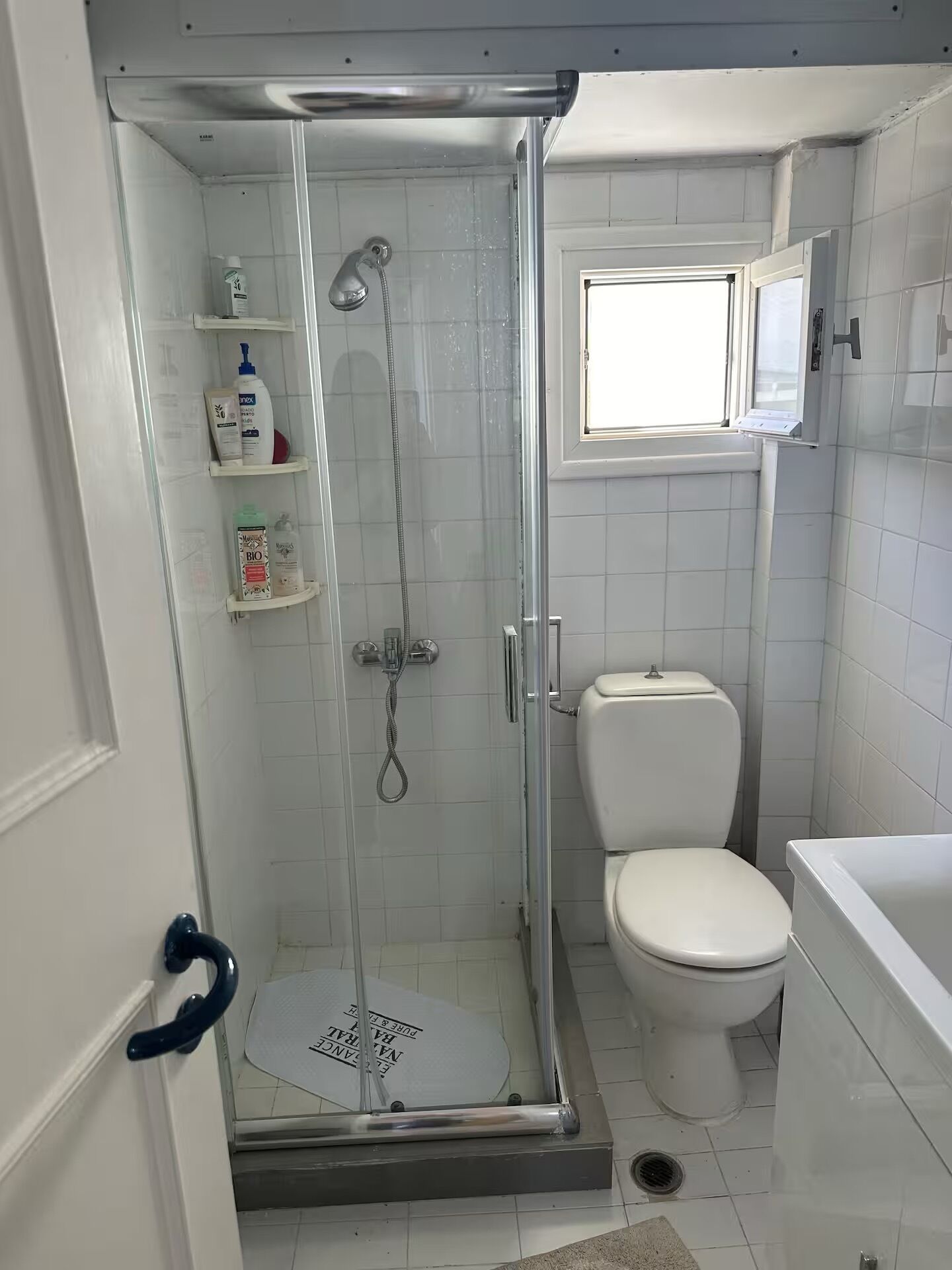 A bathroom with a glass-enclosed shower, a toilet, and a window above the sink.