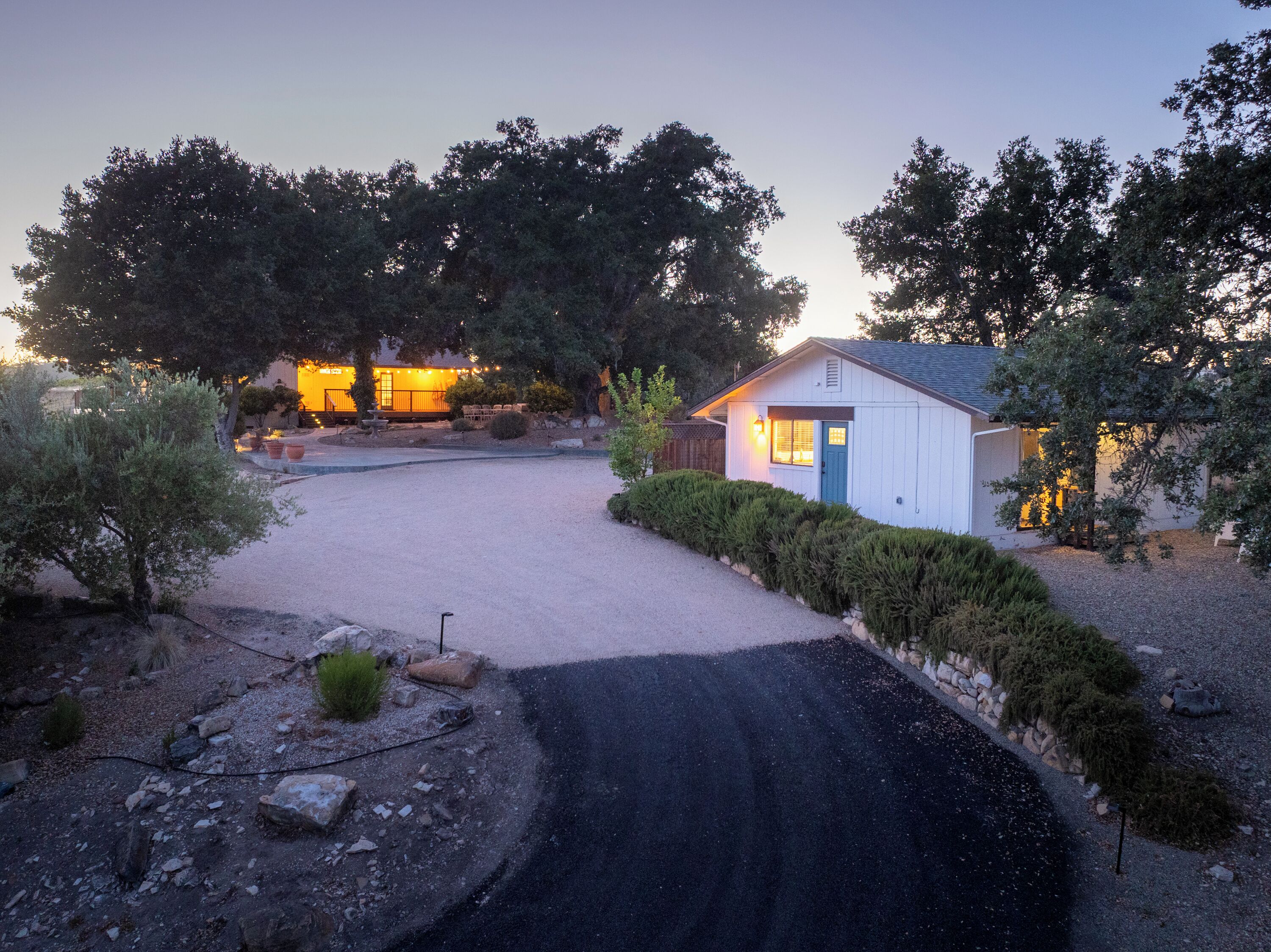 A gravel driveway leading to a small white building with a blue door, surrounded by trees and a well-maintained garden.