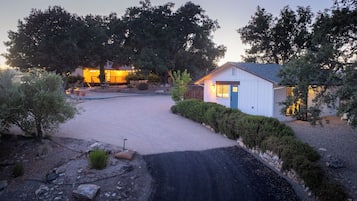 A gravel driveway leading to a small white building with a blue door, surrounded by trees and a well-maintained garden.