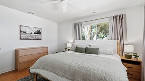 Una camera da letto con un letto, comodini, una ventola a soffitto, una finestra con tende, un quadro appeso al muro e una testiera in legno.
