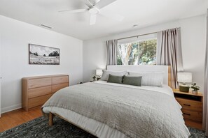 Ein Schlafzimmer mit einem Bett, Nachttischen, einem Deckenventilator, einem Fenster mit Vorhängen, einem Wandbild und einem Holz-Kopfteil.