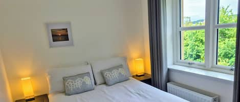 A bedroom with a bed, two bedside tables, a night lamp, a framed picture on the wall, and a window with a view of greenery.