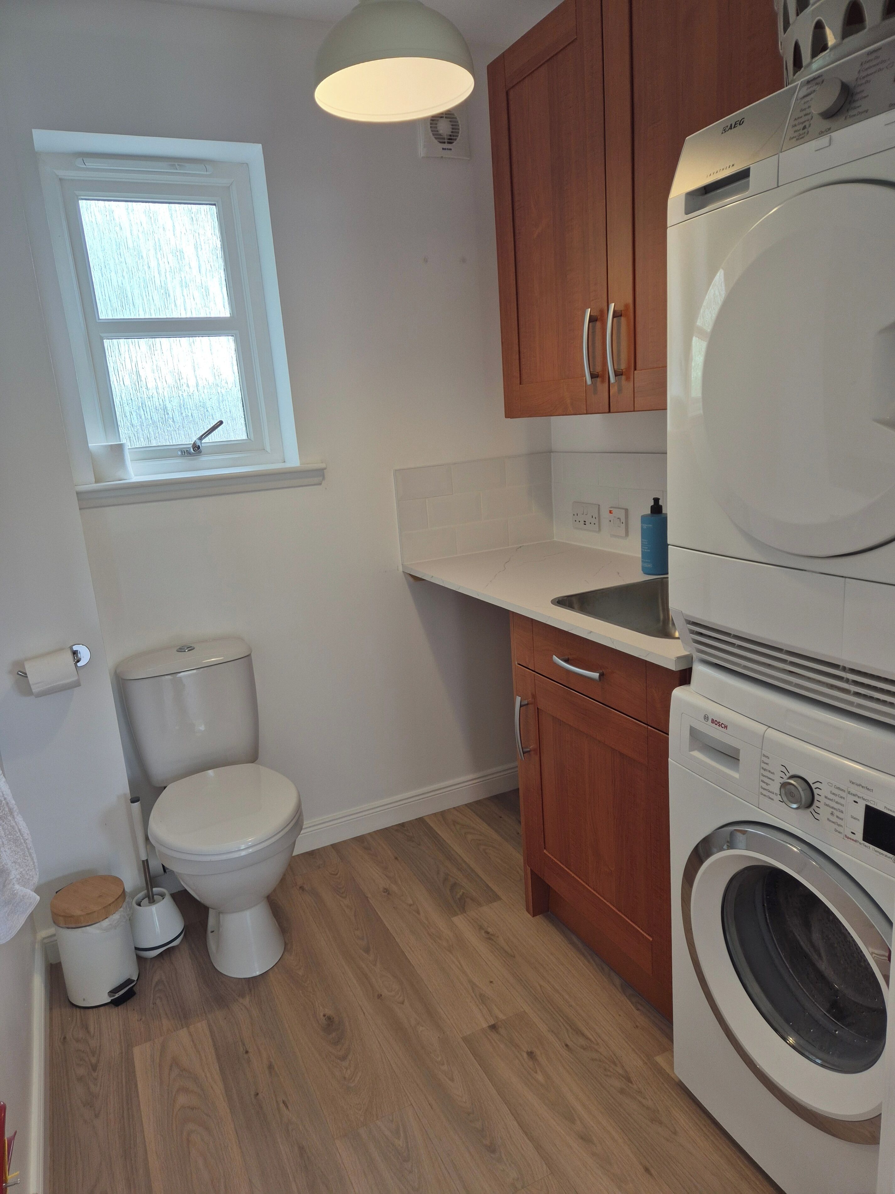 A compact bathroom with a toilet, a washing machine, wooden cabinets, and a window.
