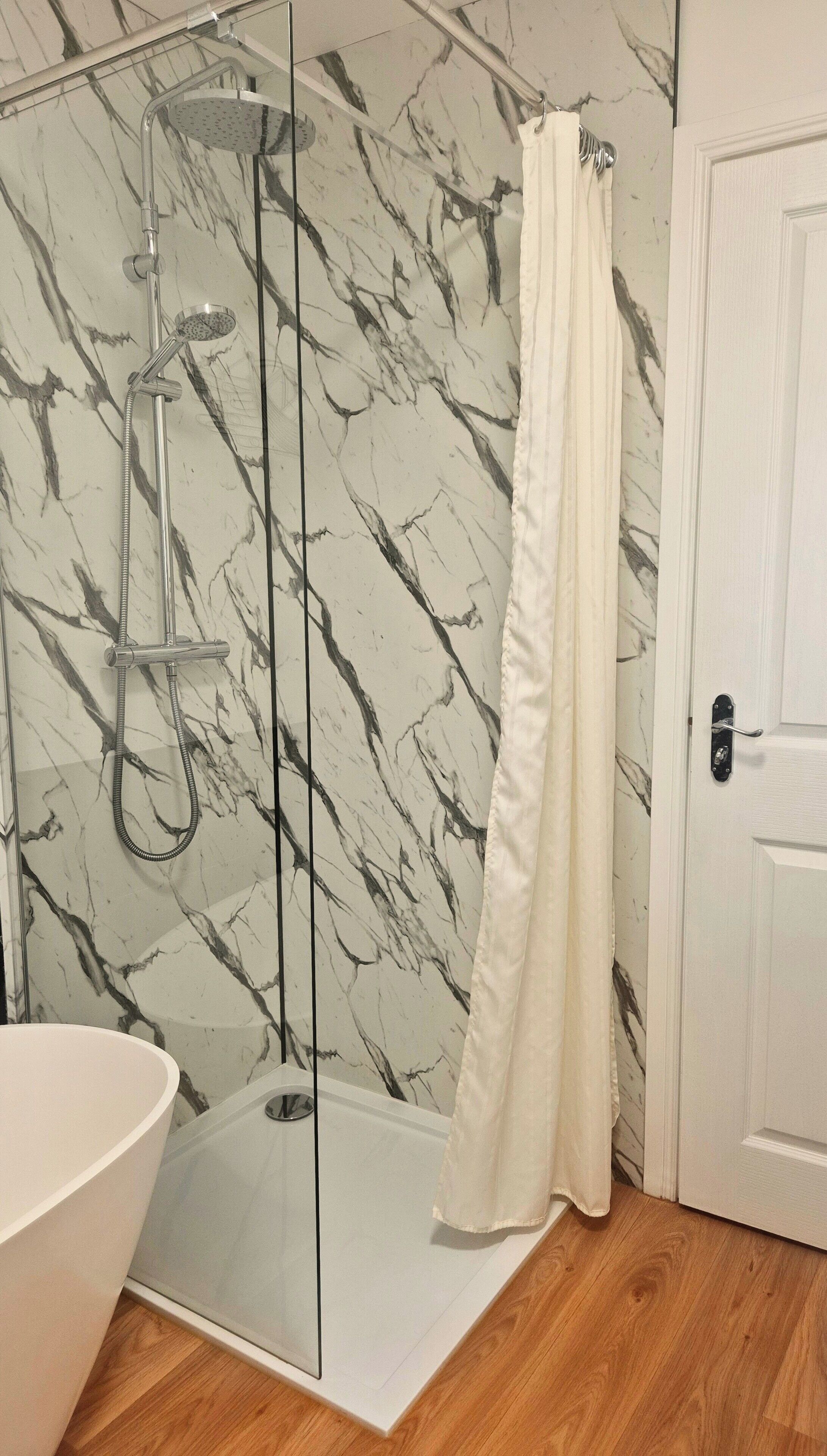 Un baño con ducha de vidrio, una bañera blanca y un revestimiento de pared con diseño de mármol.