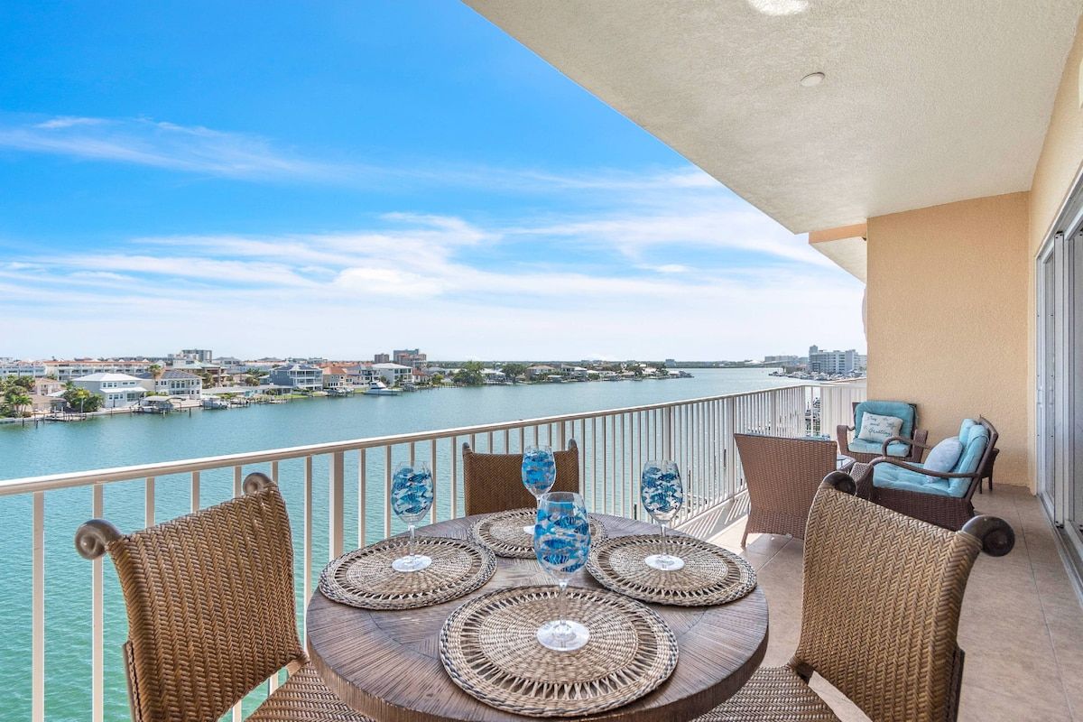 A balcony with wicker furniture, a table with glassware, and a view of a body of water and distant buildings.