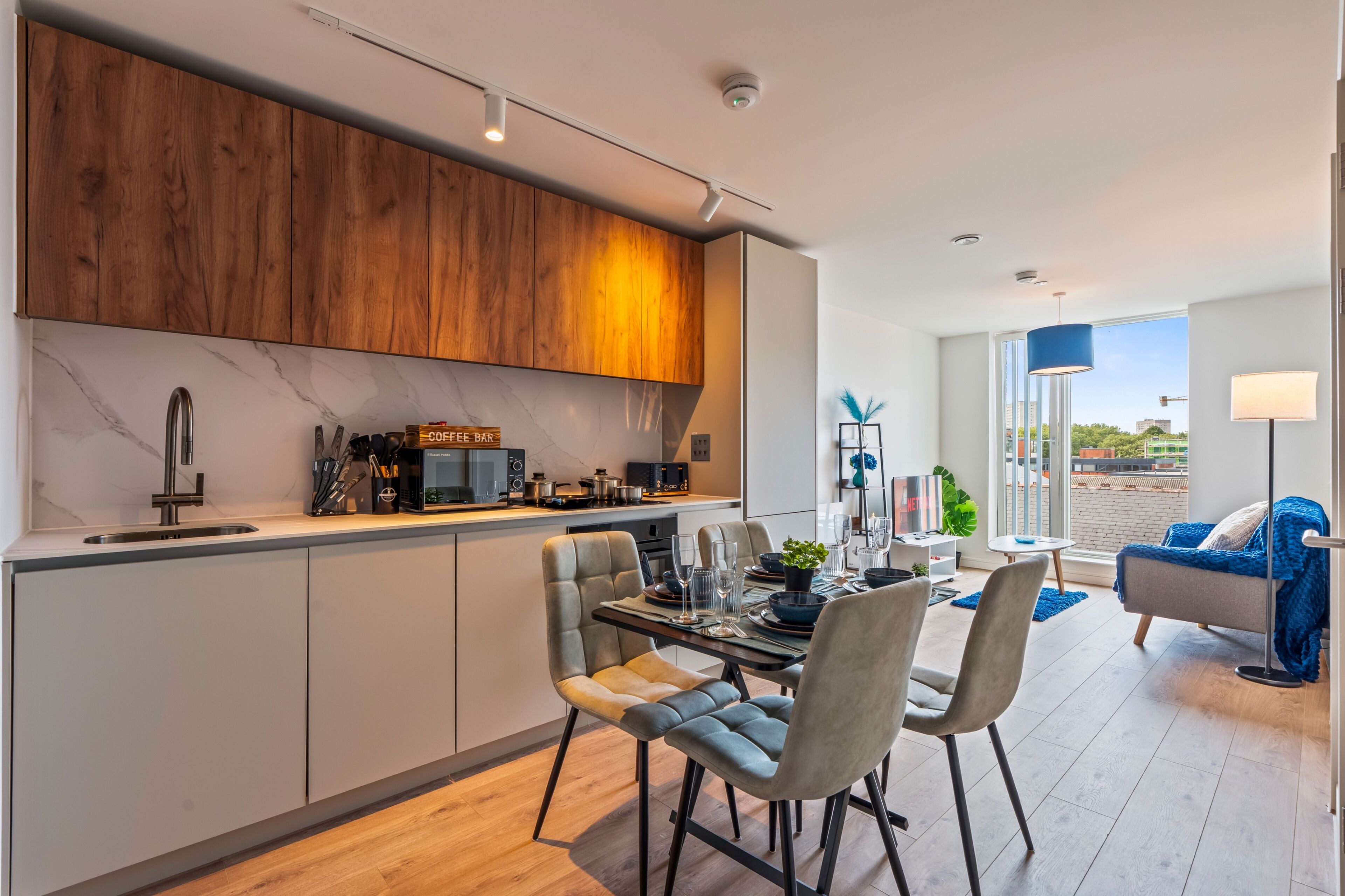 A modern kitchen with a built-in coffee bar, dining area with chairs and a table, and a living room with a sofa and a TV.