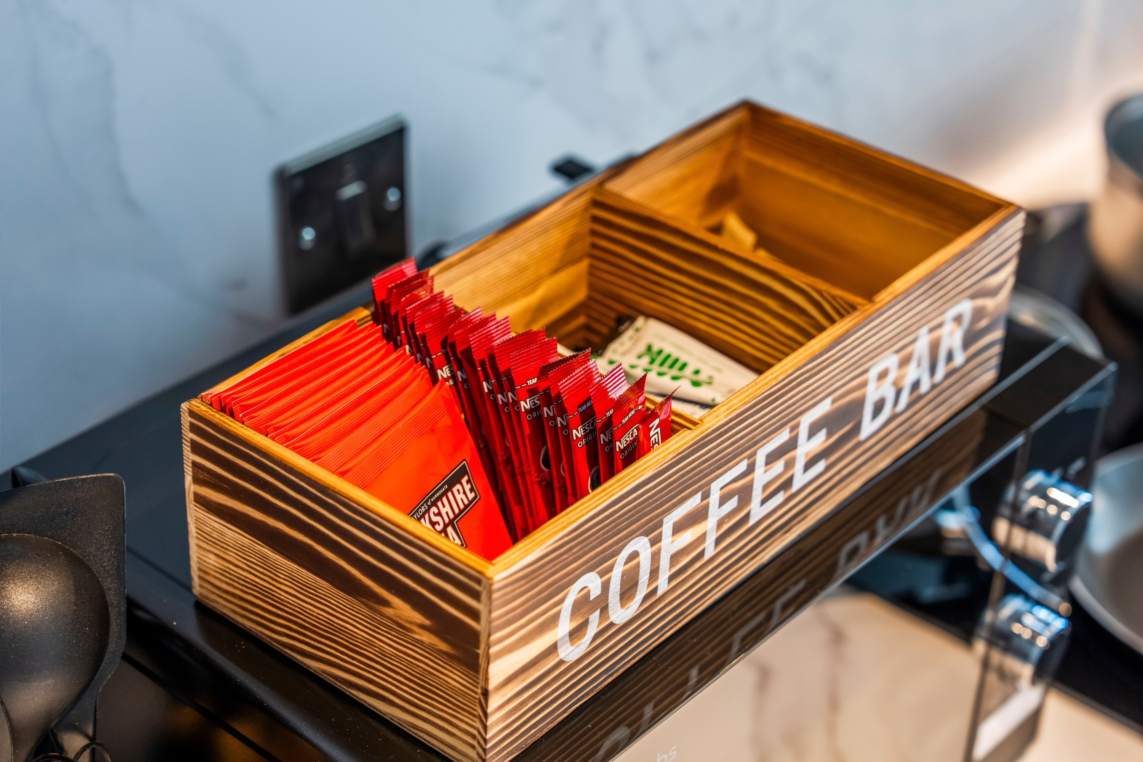 A wooden box labeled 'COFFEE BAR' containing red and white coffee packets.