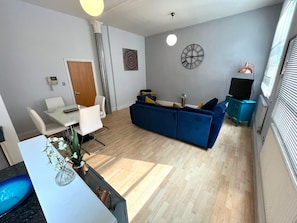 A modern living room with a sofa, a dining table with chairs, a clock on the wall, and a window with blinds.