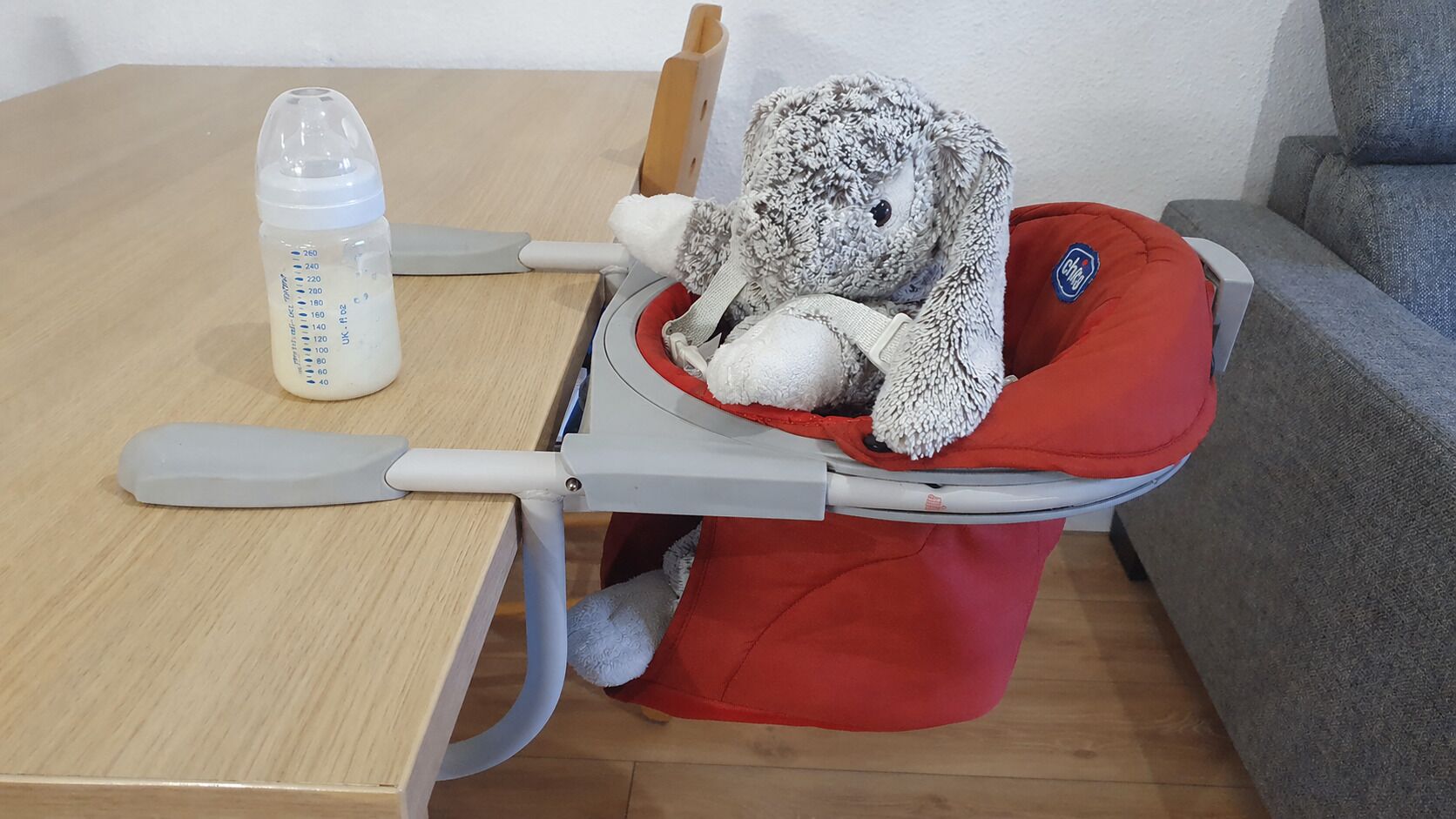 Ein Hochstuhl mit rotem Kissen, einem Stofftier, einer Milchflasche und einem Holztisch.