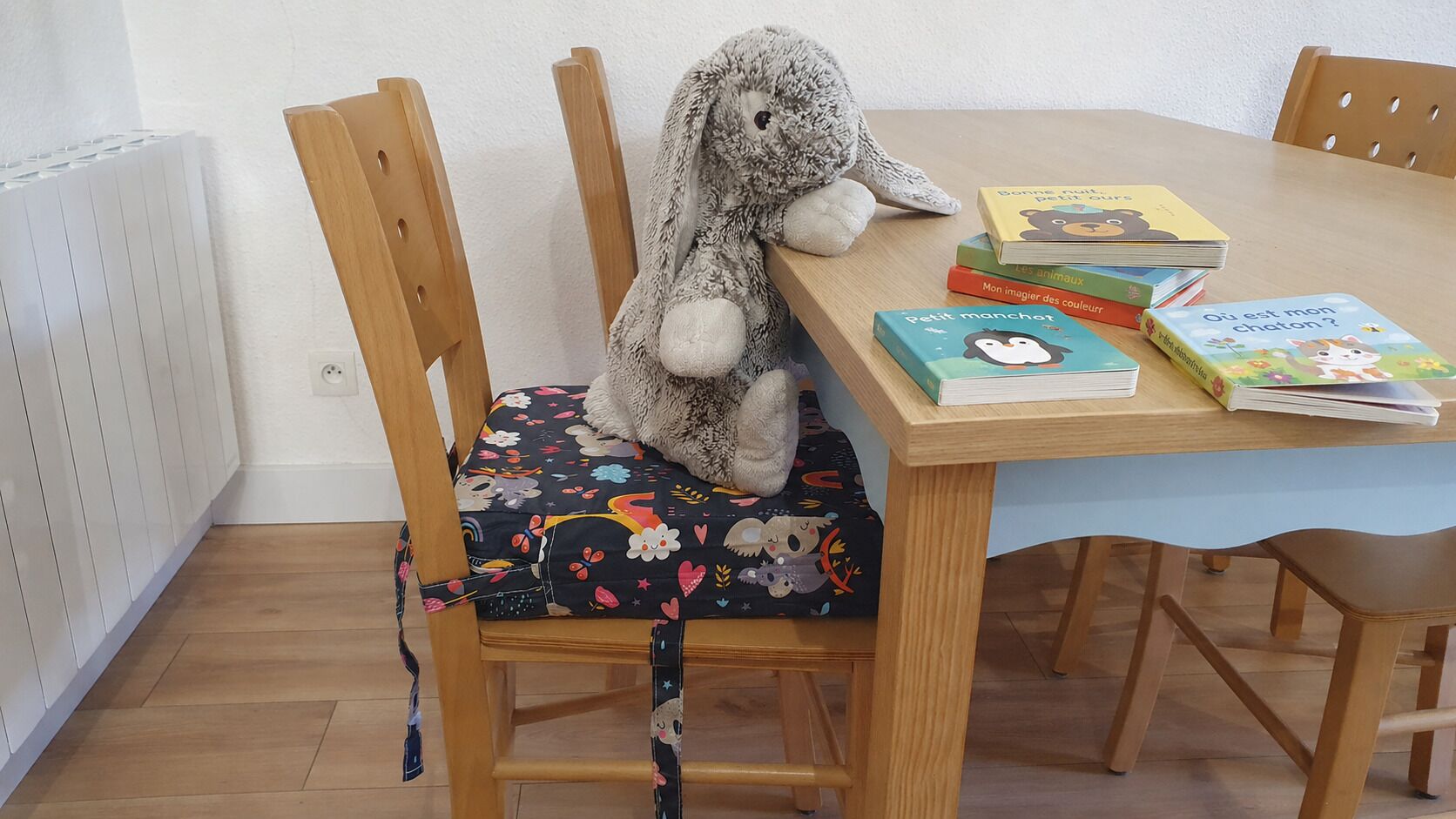 Un coin repas avec une table en bois, une chaise dotée d’un coussin à motifs floraux et un jouet en peluche.