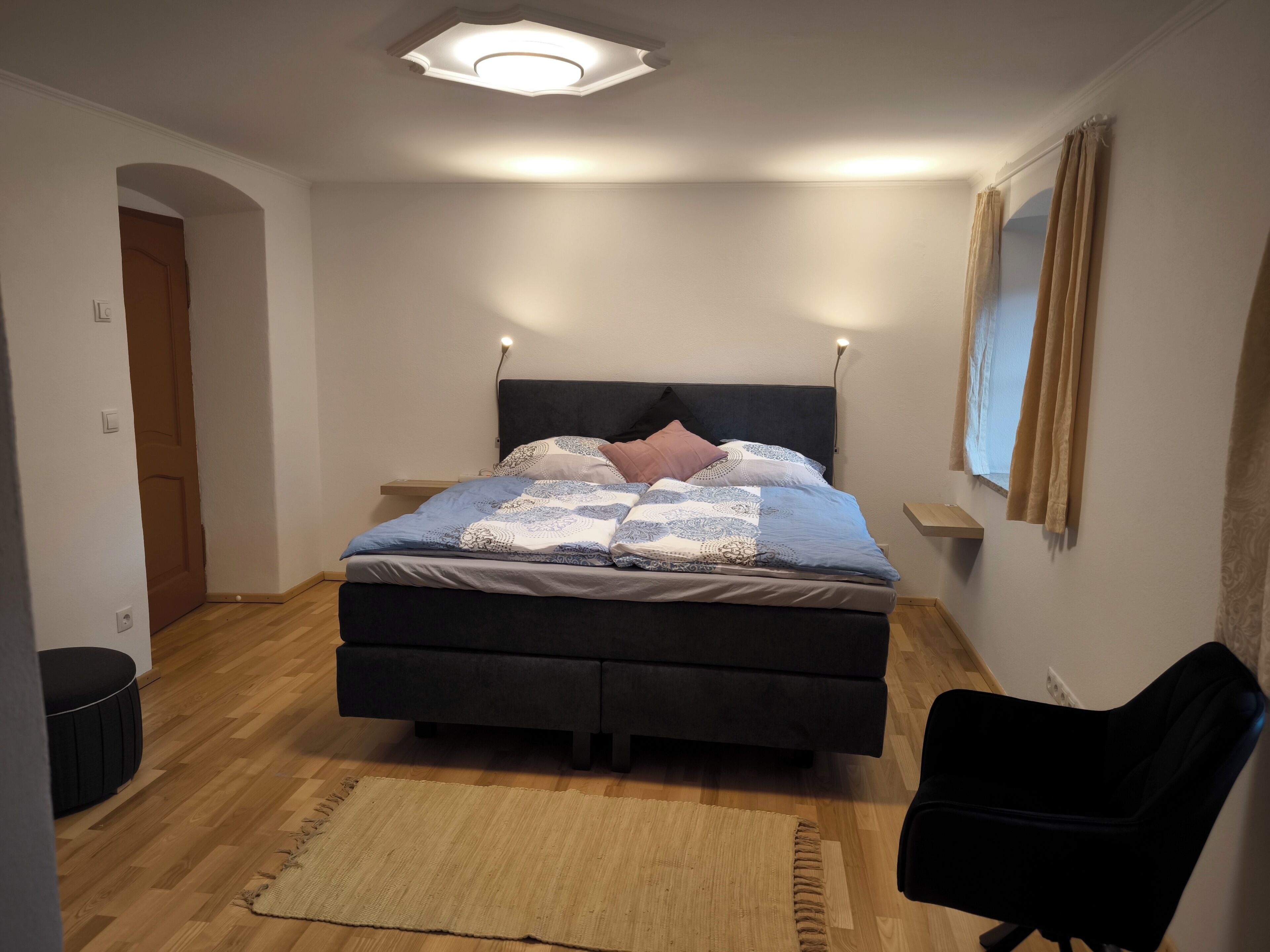 Ein Schlafzimmer mit Bett, Sessel, Teppich, Nachttisch und einem Fenster mit Vorhängen.