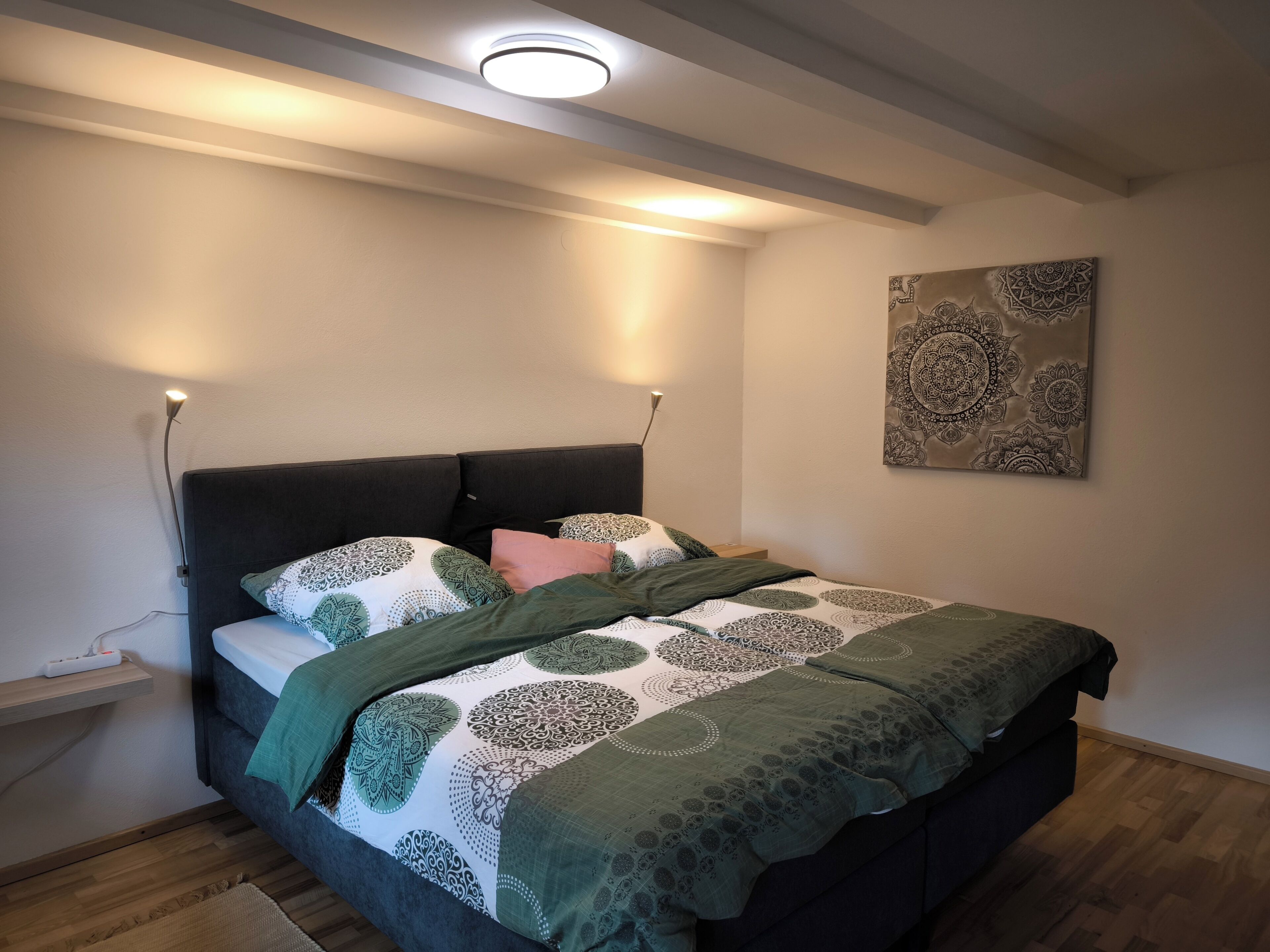 Een slaapkamer met een bed, nachtlampjes, een kunstwerk aan de muur en een stopcontact.