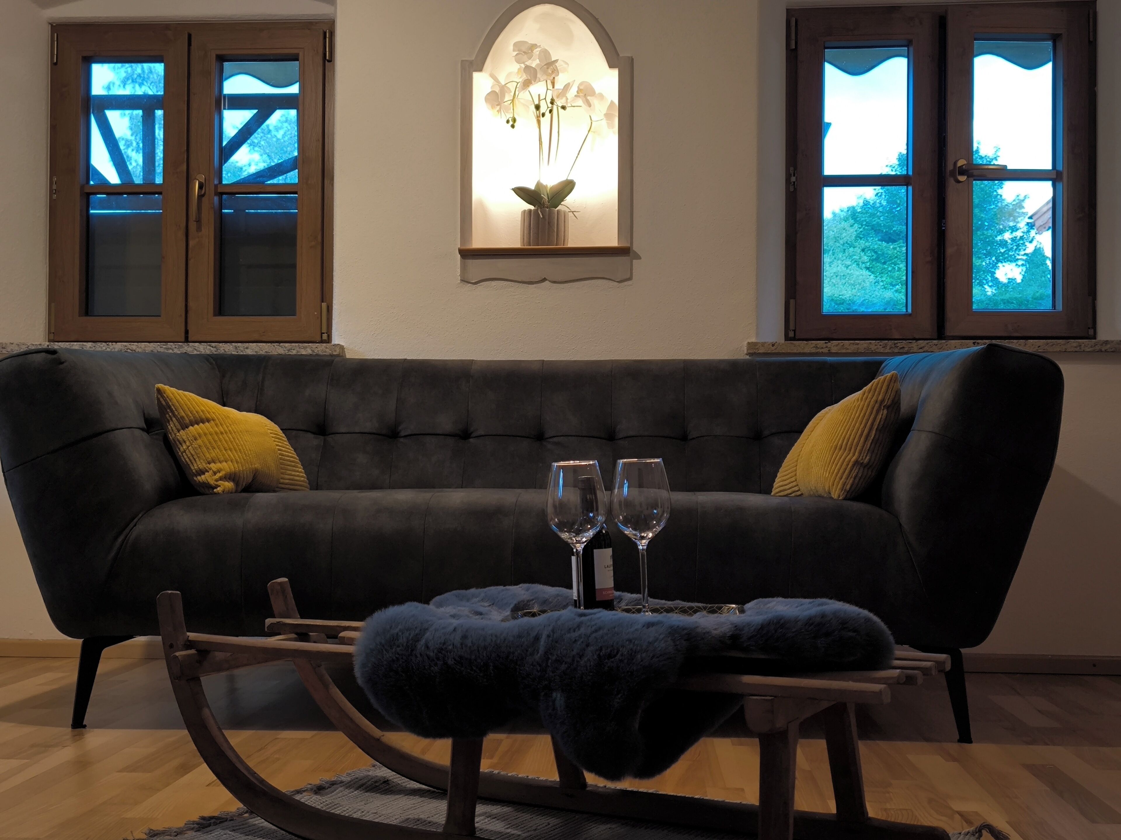 A dark-colored sofa with two yellow pillows, a small table with two wine glasses, and a decorative wall piece above the sofa.