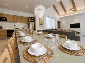 A modern dining area with a long table set for a meal, a kitchen in the background, and a living area with a sofa and a flat-screen TV. - Kamala Grand Estate 5BR Pool Villa (Tambon Kamala)