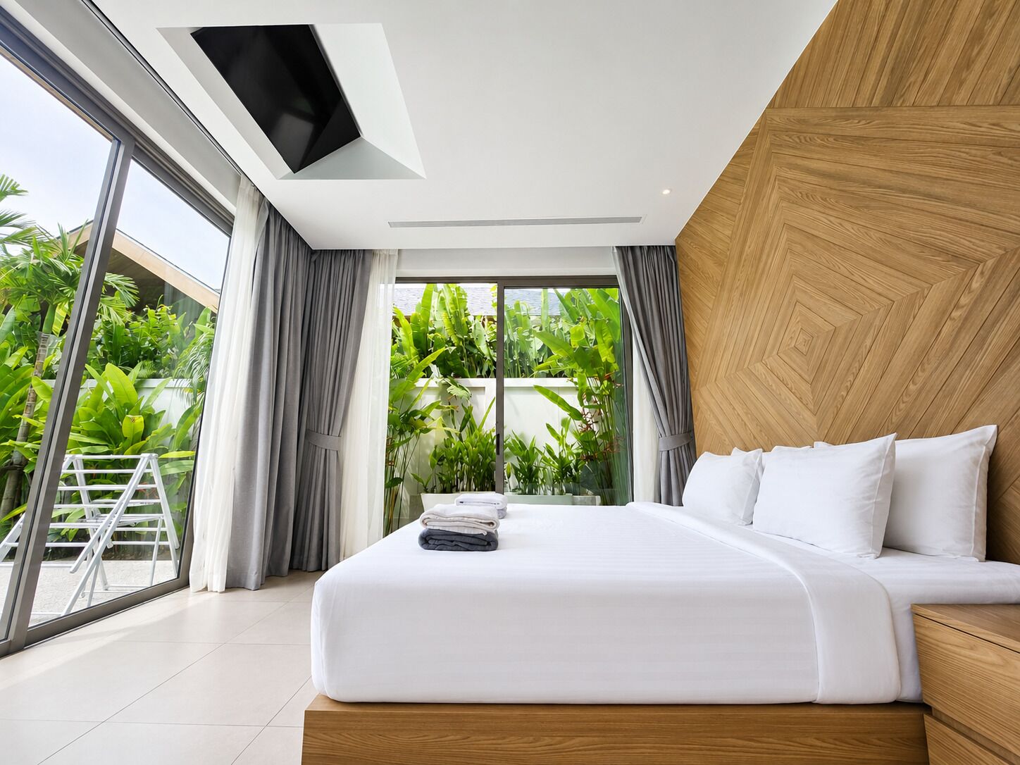 A modern bedroom with a large bed, a TV mounted on the ceiling, and a view of lush greenery outside.
